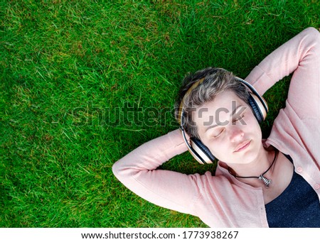 Woman in pink coat, black top and jeans listens to music / audiobook and resting on meadow / green grass on sunny spring warm day in park