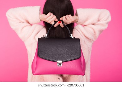 Woman in a pink coat holding handbag with two hands behind the back on a pink background