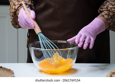 Woman in pink gloves and a brown apron beats yellow eggs with a metal whisk in a glass bowl