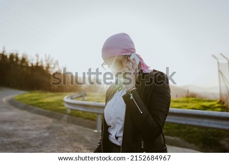 Woman in pink headscarf and sunglasses talking on smartphone while standing on path on street of city on sunny summer day
