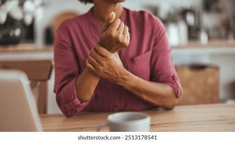 A woman in a pink shirt sits at a table, holding her wrist in discomfort, symbolizing wrist pain or arthritis in a home setting. - Powered by Shutterstock