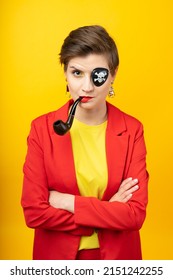 A woman with a pirate patch over one eye smokes a pipe. A sly expression on his face. Short haircut, red jacket, yellow T-shirt, bright yellow background in the studio, earrings. Modern pirate girl