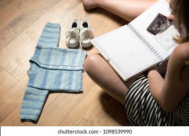 Woman Placing Baby's Sonogram Into Baby's First Year Memory Book. Baby Clothes And Sneakers Laying On The Floor