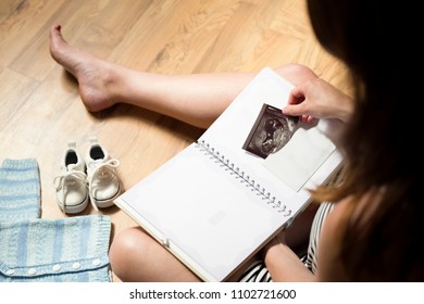 Woman Placing Baby's Sonogram Into Baby's First Year Memory Book. Baby Clothes And Sneakers Laying On The Floor