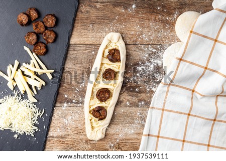 a woman is placing ingredients on flattened dough on wood kitchen countertop. A bread making concept image with loafs,  cheese and sucuk in back. Turkish kasarli sucuklu pide recipe