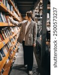 A woman in a plaid blazer reaches for a book on a shelf filled with many other books, browsing in a bookstore.