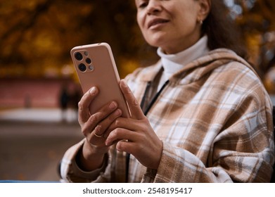 A woman in a plaid jacket enjoys browsing on her smartphone during a pleasant autumn day. The blurred background of fall colors and outdoor setting adds warmth to the scene. - Powered by Shutterstock