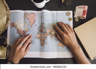 Woman Planning Her Trip. Human Hands On The World Map With The Hat, Film Camera, Some Money, Notebook From Recycled Paper And Freshly Brewed Coffee Cup On The Dark Wooden Table Background.