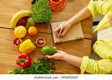 Woman Planning, Writing Weekly Meals On A Meal Planner Note Or Diet Plan On Wooden Table With Healthy Food Fruit And Vegetables In Her Kitchen At Home.