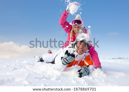 Woman playfully attacking her boyfriend with a big snowball while lying on his back 