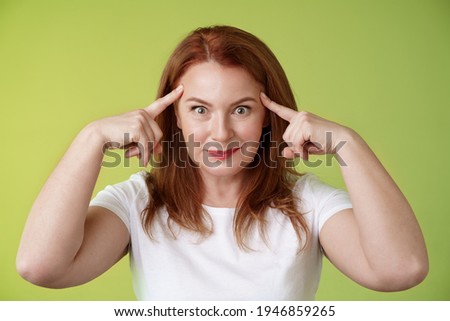 Woman playfully staring funny control your mind. Silly redhead middle-aged female touch temples popping eyes smiling delighted read thoughts trying guess intrigued what thinking green background