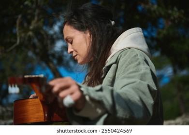 A woman playing an acoustic guitar outdoors, enjoying a tranquil moment in nature. The setting is serene, capturing the essence of music and relaxation. - Powered by Shutterstock