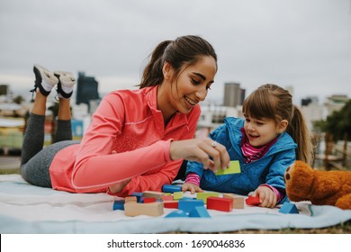 Woman playing building blocks with a small girl at the park. Girl child and her nanny lying on the blanket and playing wooden blocks at the park.