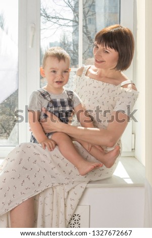 Woman is playing with the cute baby boy and reading a book to him sitting by the window with bouquet of rose tulips. Spring, new Life, tenderness conception.