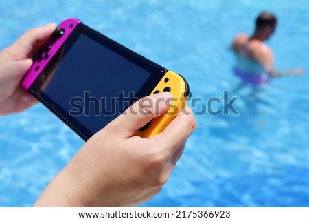 Woman playing game on handheld console near the swimming pool, selective focus on hand. Summer leisure on a beach
