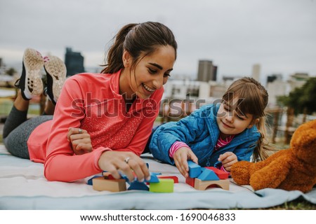 Woman playing with a small girl at the park. Girl child playing building blocks with her nanny at the park. Both lying on a plaid.