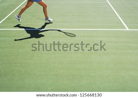 woman playing tennis, only half of her body visible, shadow holding a tennis racket seen on the ground,