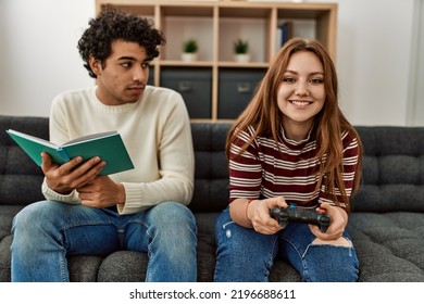 Woman Playing Video Game While Unhappy Boyfriend Read Book At Home.