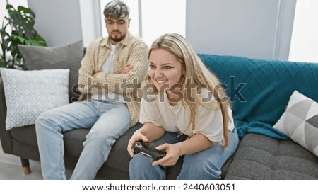 A woman plays a video game while a man watches sullenly in a bright living room, highlighting leisure, gender dynamics, and home life.