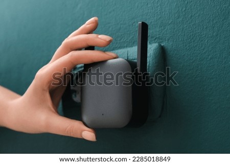Woman plugging black WiFi repeater in electric socket on green wall