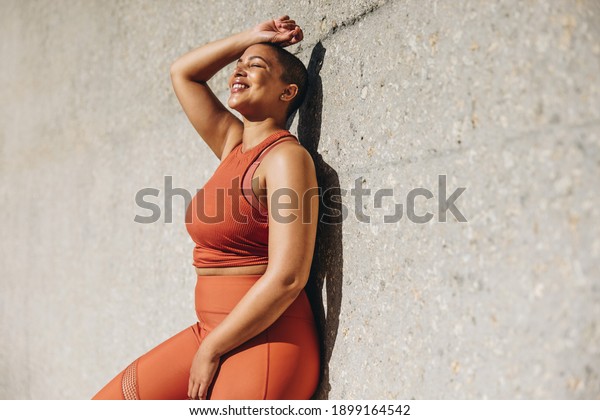 Woman with plus size body leaning to a wall and\
relaxing after workout session outdoors. Woman in sports clothing\
taking a break from\
exercise.