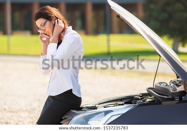 A woman plus size in a white\
shirt is standing near the open hood of her car. Waiting for help\
tow truck or technical support. A woman calls the service\
center