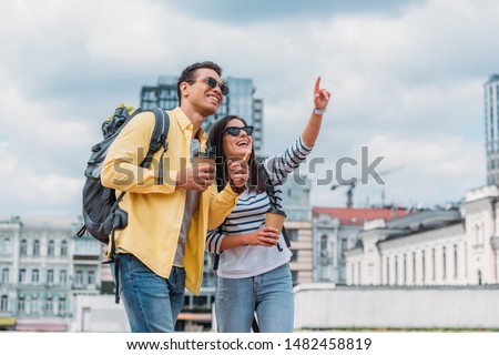 Woman pointing with finger and looking away near smiling bi-racial friend with backpack 