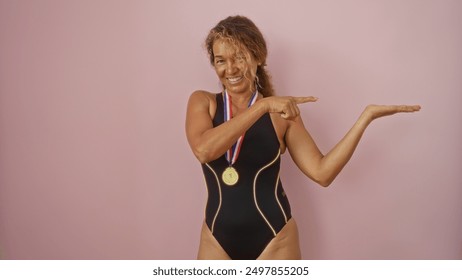 Woman pointing at palm with gold medal, wearing black swimsuit against isolated pink background - Powered by Shutterstock