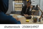 A woman police officer engages in conversation on the phone while examining documents in an office, as a man in a hoodie looks on.