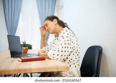 A woman in a polka dot blouse appears stressed while working remotely on her laptop in a home office environment. She holds her head in frustration. - Powered by Shutterstock