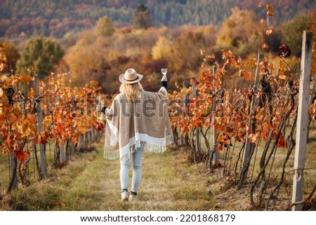 Woman with poncho and hat enjoying white wine in her vineyard at autumn. Happy vintner drinks wine after successful grape harvesting