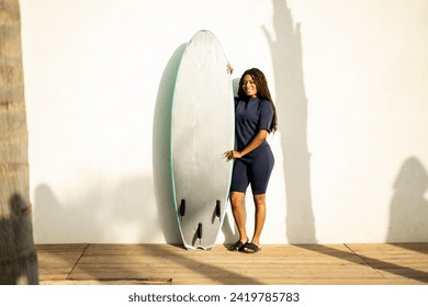 A woman poses near a wall with a surfboard dressed in a wetsuit.The African girl looks at the camera happily.Concept of selling surfboards, advertising surfing equipment. - Powered by Shutterstock