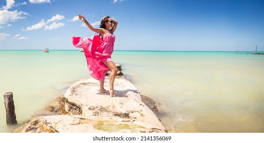 Woman posing in pink dress stands on a rock next to an exotic sea in a summer holiday destination.