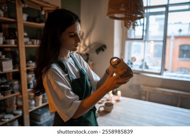 Woman potter inspects newly crafted wet clay cup in workshop for quality and shape. Craftswoman evaluate handmade pottery, checking craftsmanship, pottery refinement, ceramic artistry on master class - Powered by Shutterstock