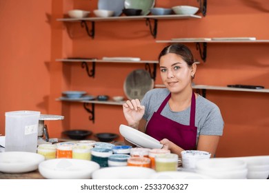 Woman potter with paintbrush glazing, painting on plate in workshop, working in pottery studio. - Powered by Shutterstock