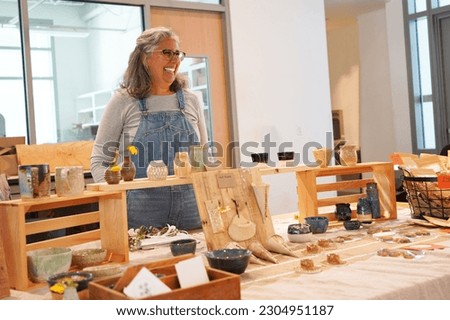 Woman pottery artist wearing overalls stands at her display table selling pottery items at a crafts fair, smiling