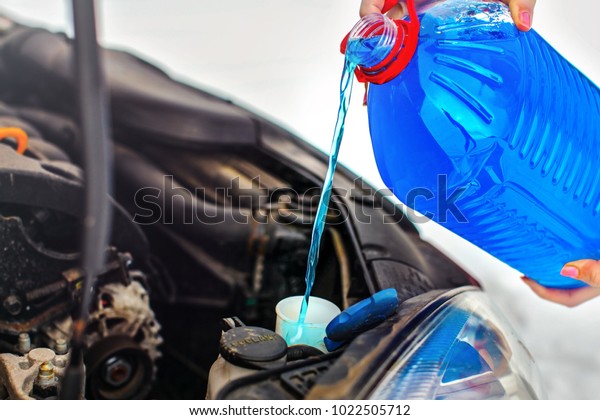 Woman pouring antifreeze car\
screen wash liquid into dirty car from blue anti freeze water\
container.