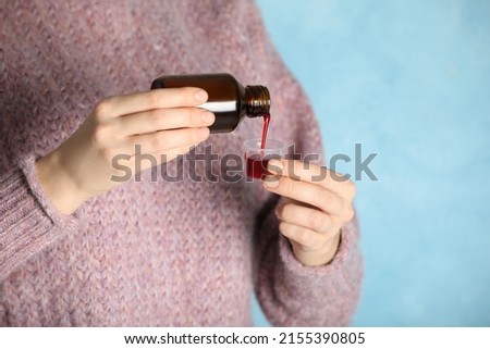 Woman pouring cough syrup into measuring cup on light blue background, closeup