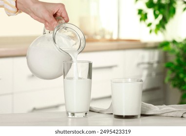 Woman pouring fresh milk from jug into glass at light wooden table in kitchen, closeup