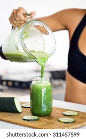 Woman Pouring A Green Detox Juice From Blender To A Jar, At Home Wearing Sport Clothes.