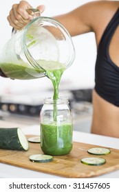 Woman Pouring A Green Detox Juice From Blender To A Jar, At Home Wearing Sport Clothes.