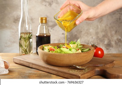Woman pouring honey mustard dressing into bowl with fresh salad on table