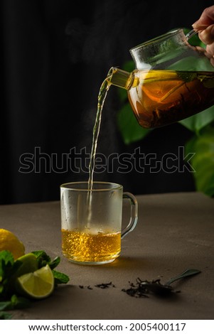 Woman pouring hot black tea with mint, lemon and lime from teapot into glass mug, evening tea party