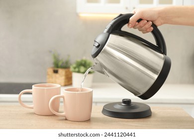 Woman pouring hot water from electric kettle into cup in kitchen, closeup