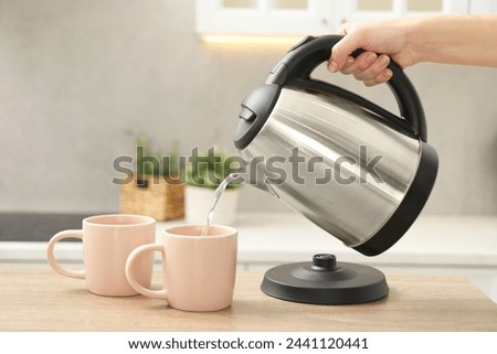 Woman pouring hot water from electric kettle into cup in kitchen, closeup