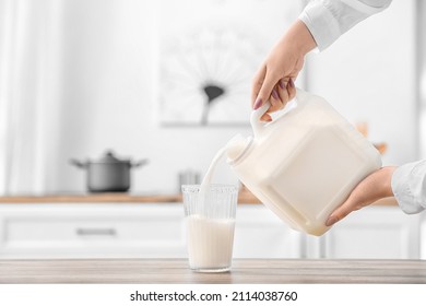 Woman Pouring Milk From Gallon Container Into Glass On Table In Kitchen