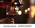 Woman pouring red wine from bottle into glass on blurred background, closeup. Space for text