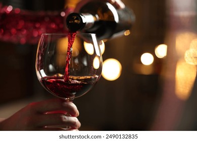 Woman pouring red wine from bottle into glass on blurred background, closeup. Space for text