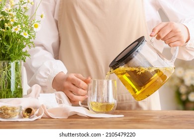 Woman pouring tasty chamomile tea from teapot into cup on table