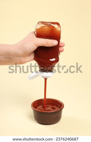 Woman pouring tasty ketchup from bottle into bowl on beige background, closeup
